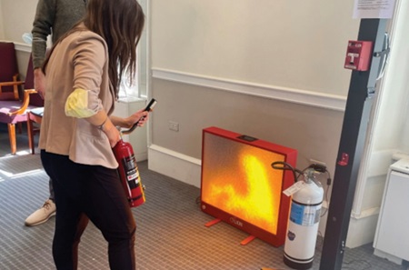 An employee holds a mock fire extinguisher and holds it towards a small screen with virtual flames